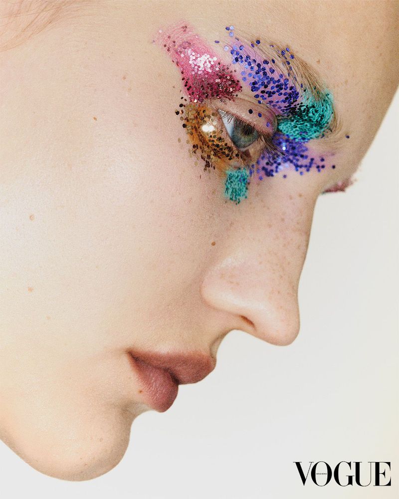 Extreme close-up on a fair-skinned adult woman with glitter makeup around the eye, emphasizing artistic beauty and Vogue branding.