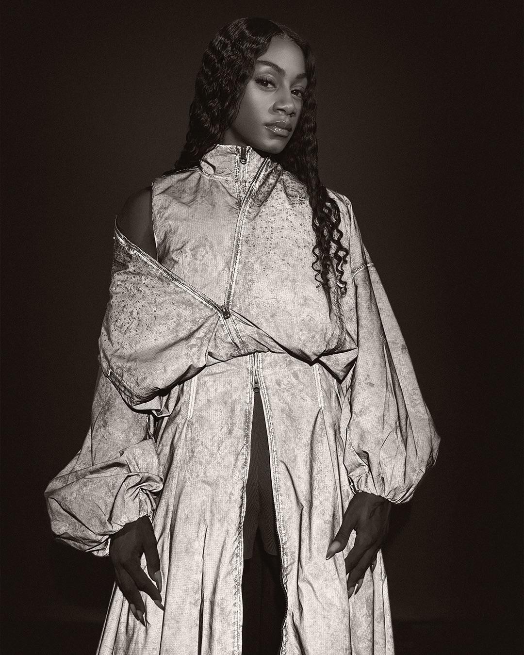 Portrait of Sha'Carri Richardson wearing a structured grey patterned jacket against a dark background for Nike Atelier.
