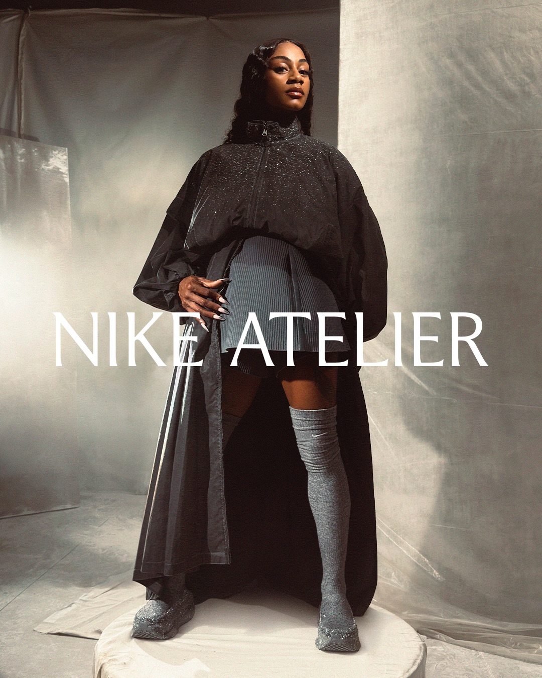 Sha'Carri Richardson wearing an oversized black coat and grey knee socks on a studio pedestal for Nike Atelier.