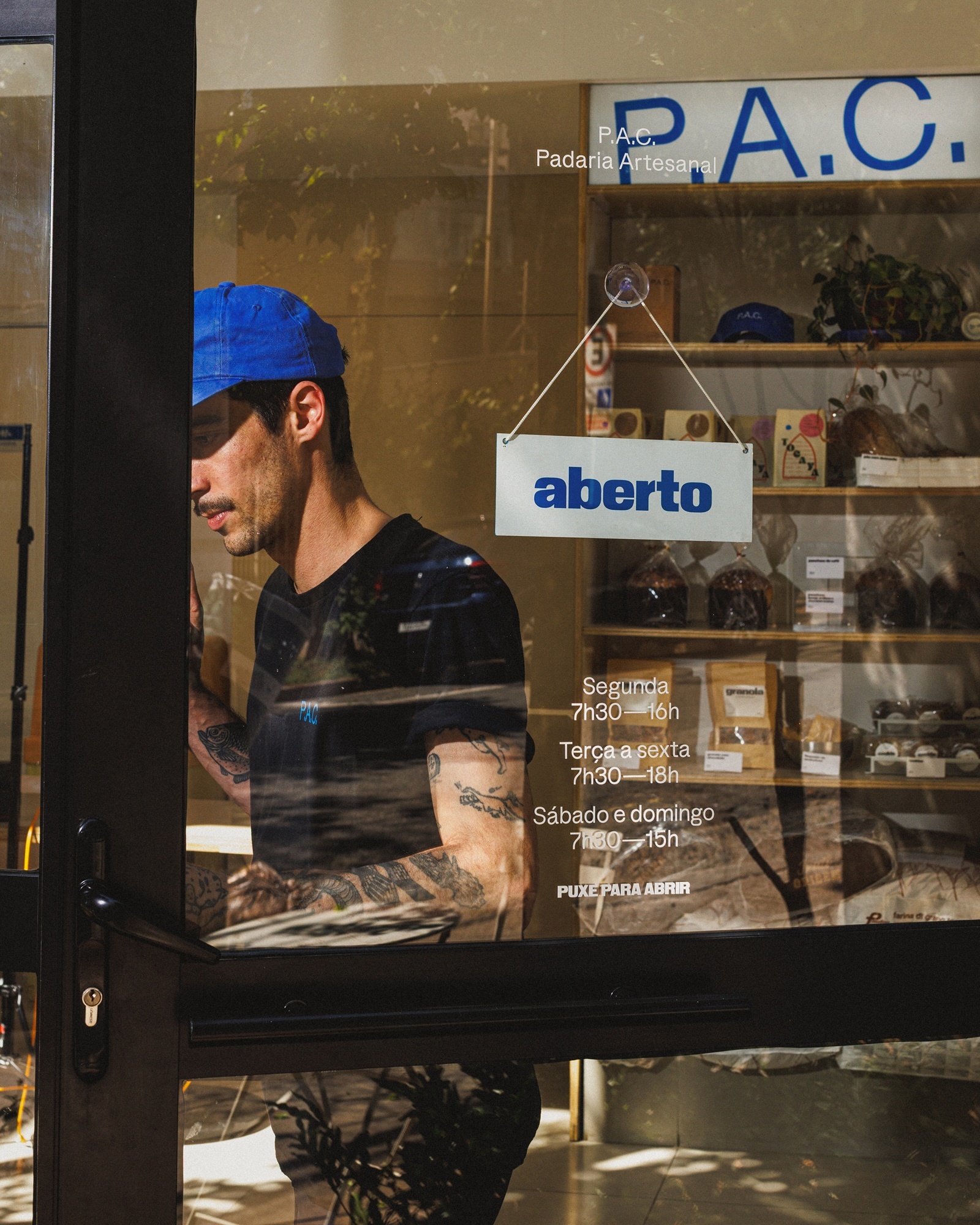 Man wearing a blue cap and black t-shirt standing behind the glass storefront of P.A.C. Padaria Artesanal