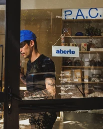 Man wearing a blue cap and black t-shirt standing behind the glass storefront of P.A.C. Padaria Artesanal