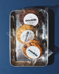 Three individually wrapped artisanal cookies inside a metal tray resting on a dark blue background.