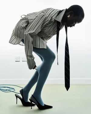 Model leaning forward against a white wall wearing a striped shirt, tie, light blue tights, and black heels.