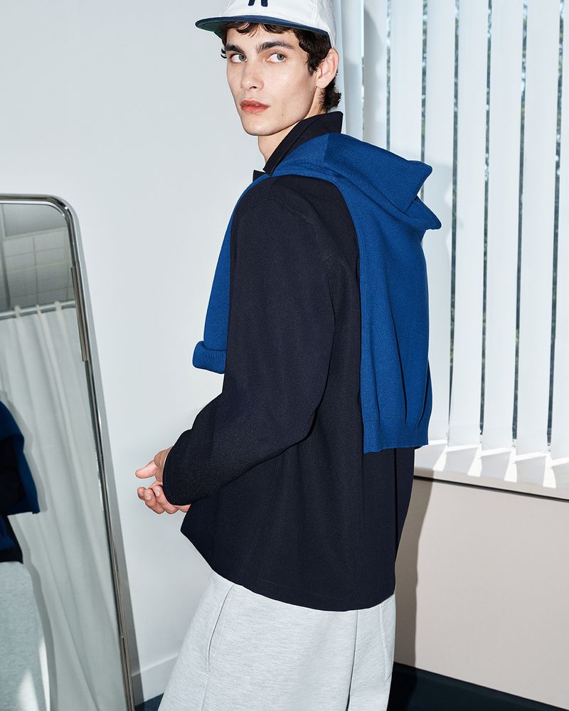 Model in white cap wearing dark jacket with blue sweater draped over shoulders and light grey trousers