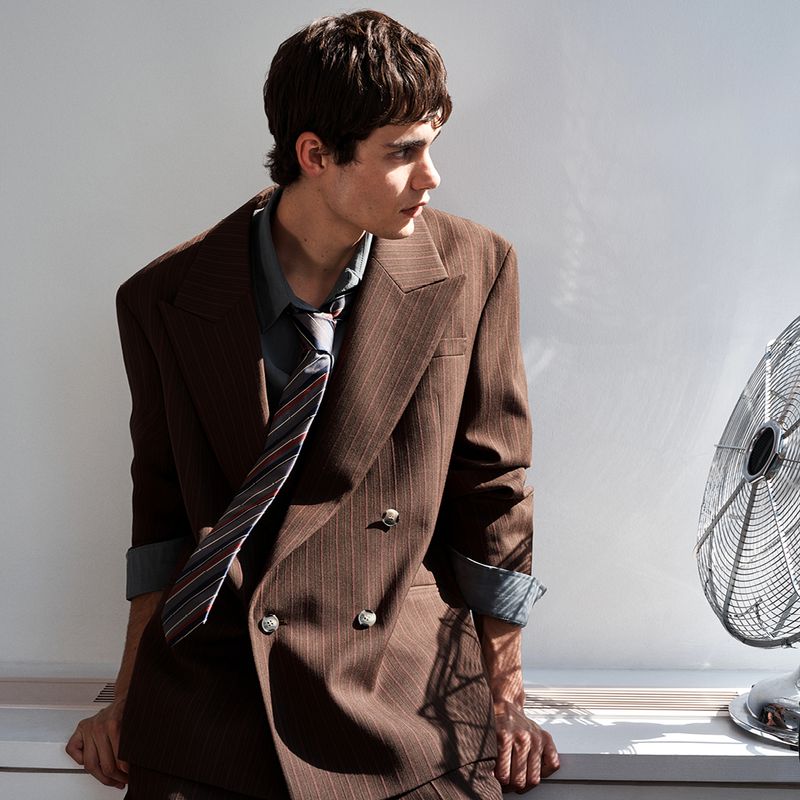 Model in a brown pinstripe double-breasted blazer and striped tie leaning near an electric fan against a plain wall.