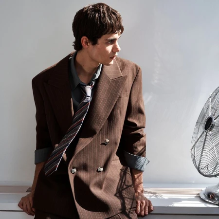 Model in a brown pinstripe double-breasted blazer and striped tie leaning near an electric fan against a plain wall.