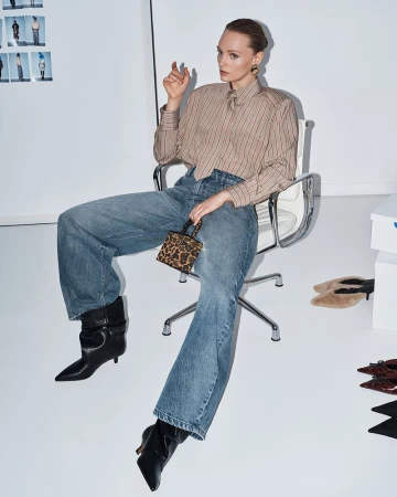 Model seated on a white office chair wearing a striped shirt, blue jeans, black boots, and holding a small animal-print handbag