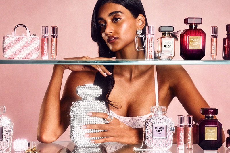 Model Neelam Gill posing behind a glass shelf displaying Victoria's Secret perfume bottles and lip glosses against a pink background.
