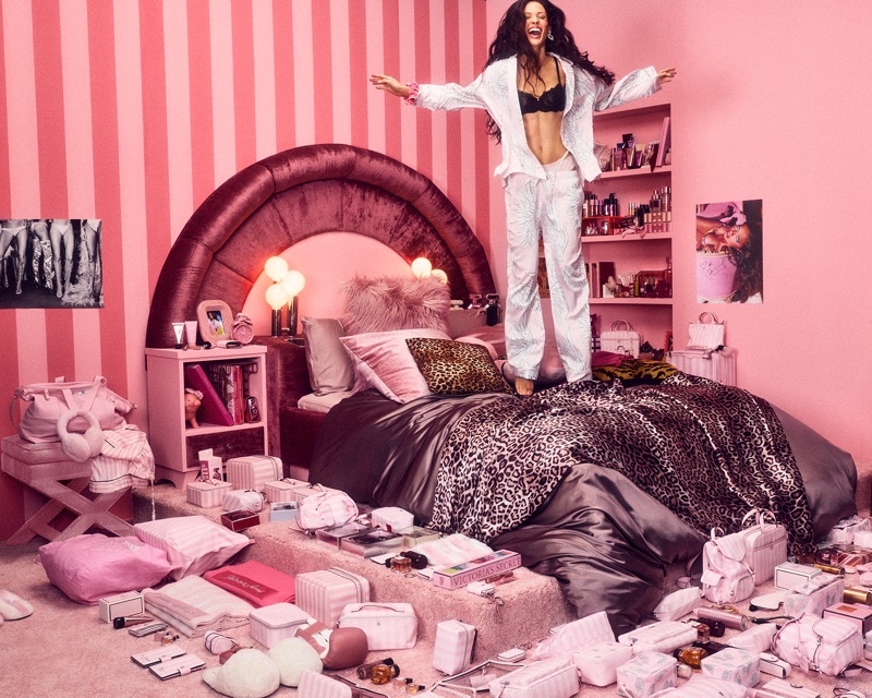 Model jumping joyfully on a bed in a pink bedroom surrounded by packaged beauty products for Victoria's Secret.
