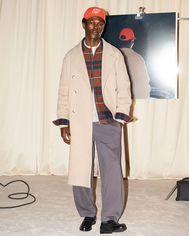Man wearing a long cream coat over a plaid shirt, grey trousers, loafers, and a red cap in a studio.