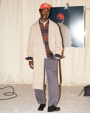 Man wearing a long cream coat over a plaid shirt, grey trousers, loafers, and a red cap in a studio.