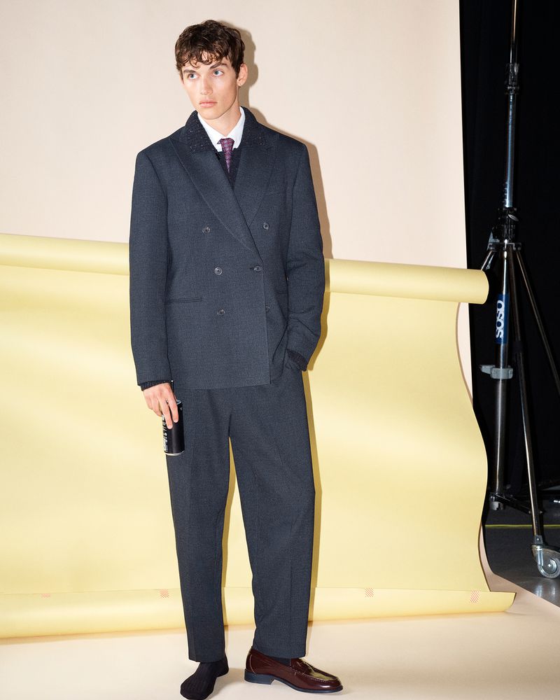 A male model wearing a dark grey double-breasted suit holding a can in a studio with a yellow backdrop roll.