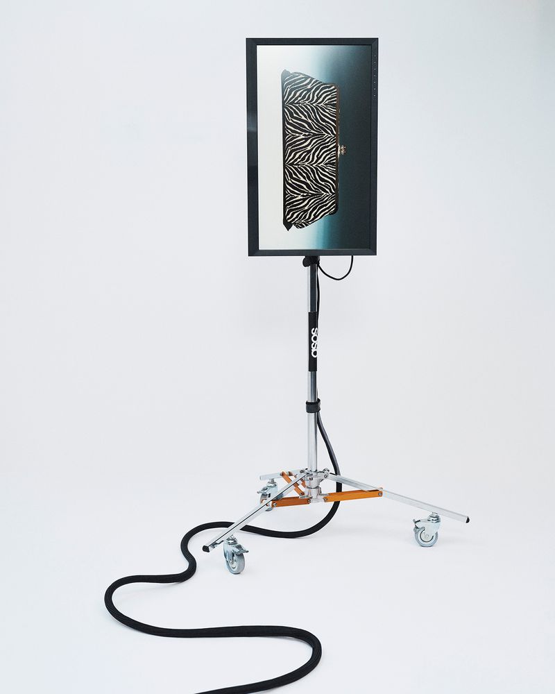 A digital screen displaying a zebra print handbag on a wheeled studio light stand against a white backdrop.