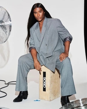 Model with long dark hair wearing a grey pinstripe suit seated on a wooden apple box in a white studio.