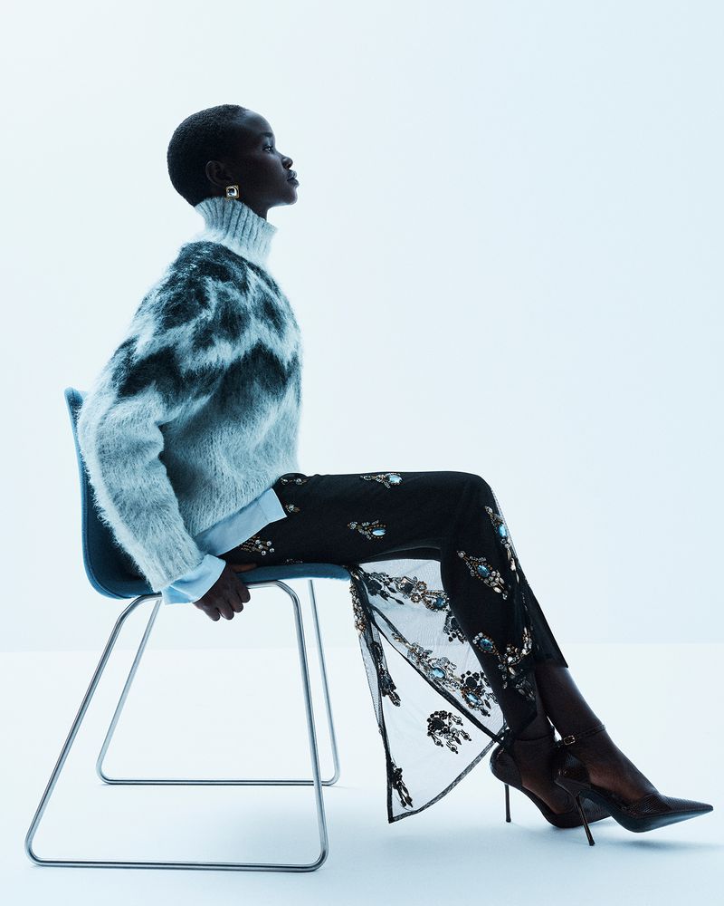 Model seated in profile on a blue chair wearing a fuzzy patterned turtleneck and embellished trousers against a white backdrop.