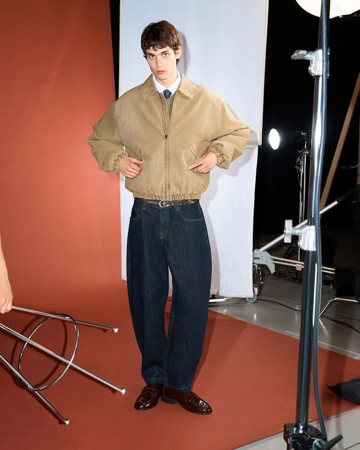 Model in a tan zip jacket and dark denim jeans standing in a photography studio with lighting equipment