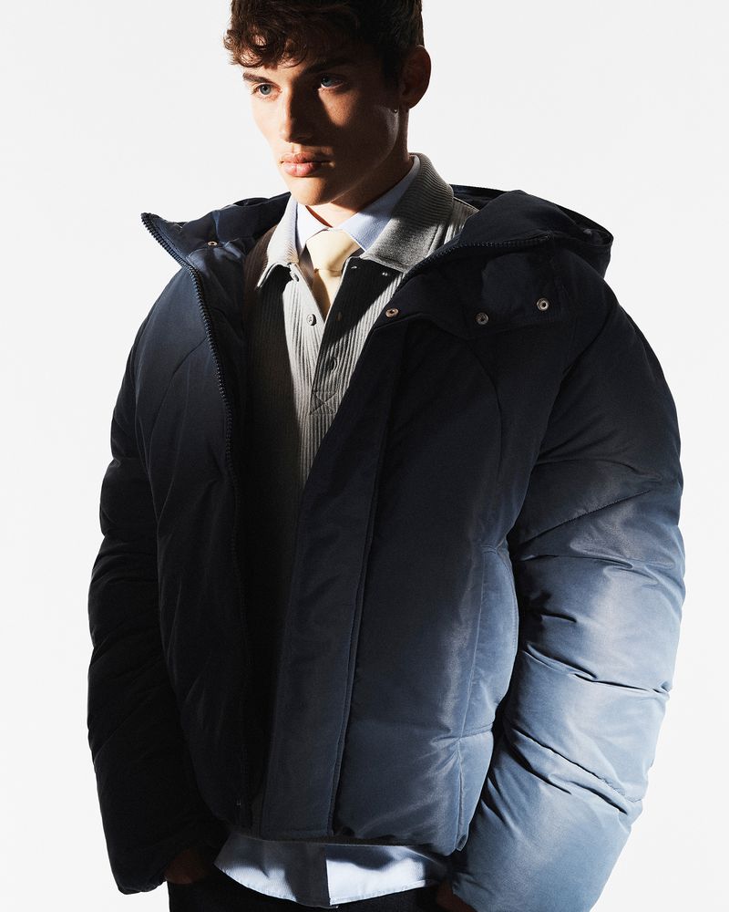 Young male model wearing a dark puffer jacket over a collared shirt against a white background