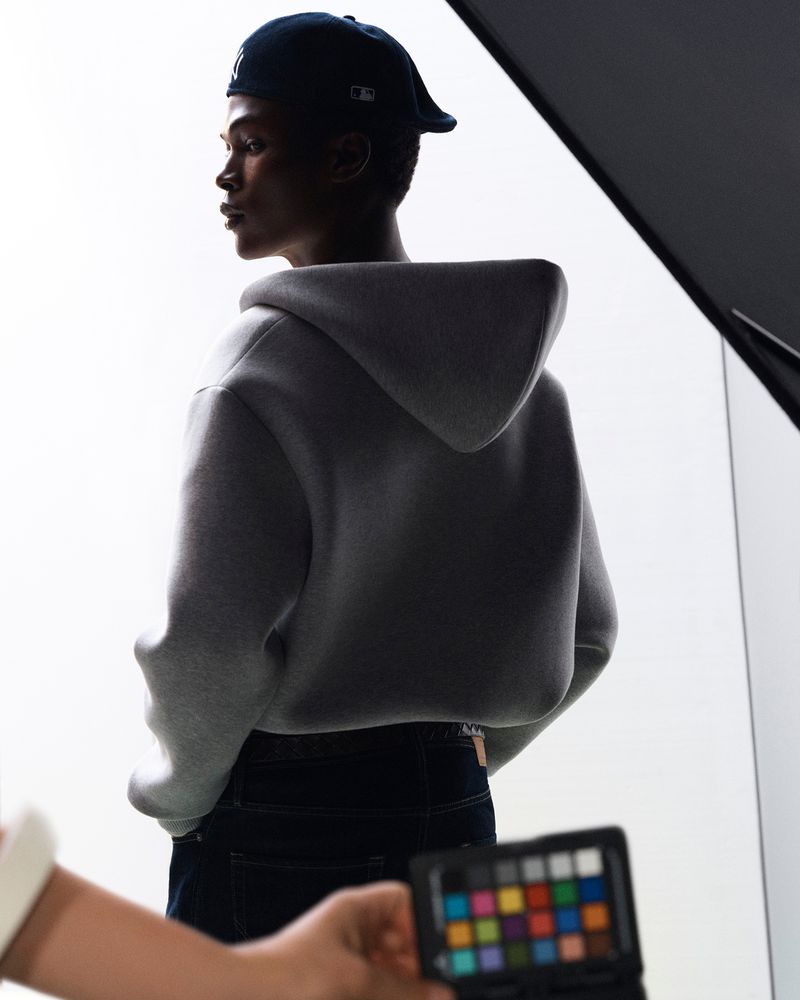 Model in grey hoodie and dark cap photographed against a bright studio background with a color chart in the foreground