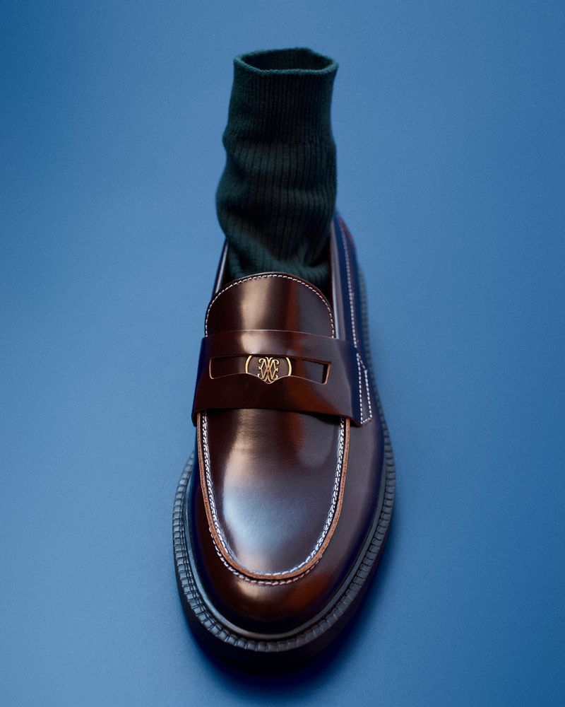 Close-up of a glossy burgundy leather penny loafer worn with a dark green ribbed sock against a blue background