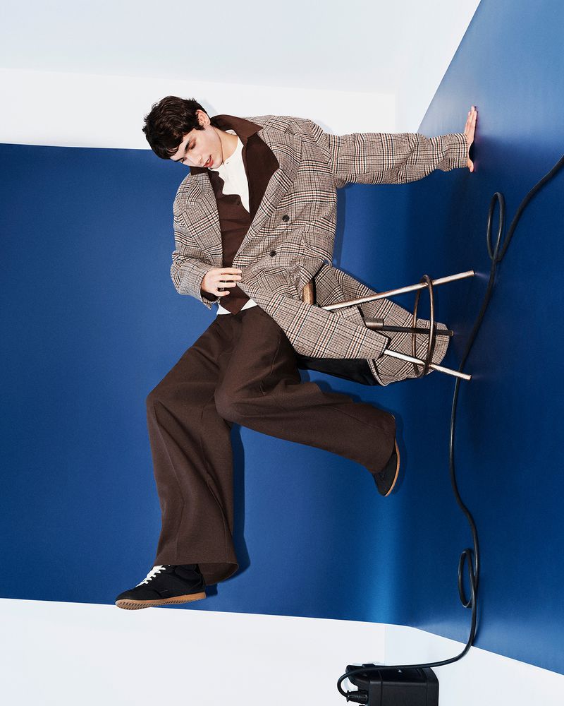 Model wearing a plaid coat and brown trousers posing against a blue and white studio wall with a stool