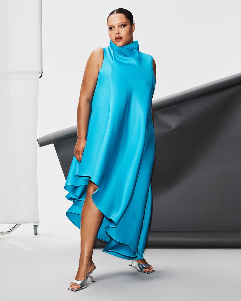 Model wearing a bright blue high-neck dress and silver mules standing against a studio background.