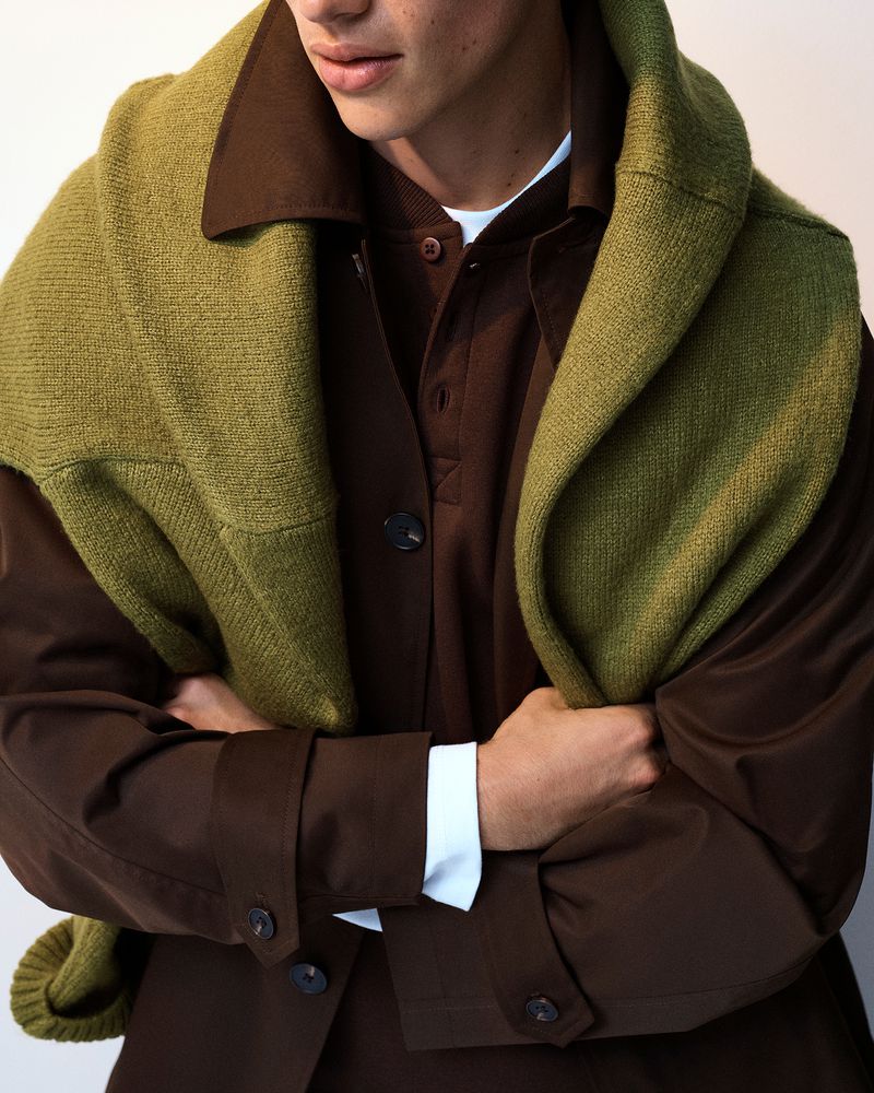 Model wearing a brown jacket with a green knitted sweater draped over the shoulders against a neutral backdrop
