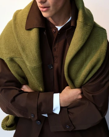 Model wearing a brown jacket with a green knitted sweater draped over the shoulders against a neutral backdrop