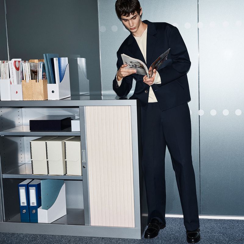 A young man in a dark suit standing beside a grey office cabinet reading a magazine