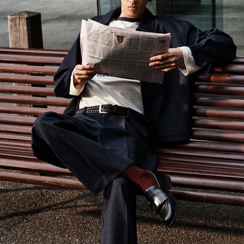 Man seated on a wooden park bench reading a newspaper, wearing a dark jacket, trousers, and loafers.