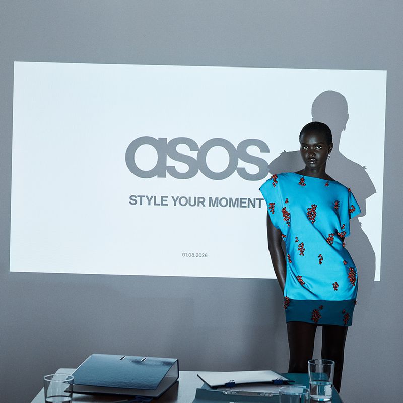 Model wearing a vibrant blue dress with dark embellishments posing in front of a wall with an ASOS logo projection