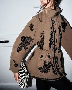 Model wearing a brown jacket with black floral embroidery holding a zebra print clutch against a plain white background