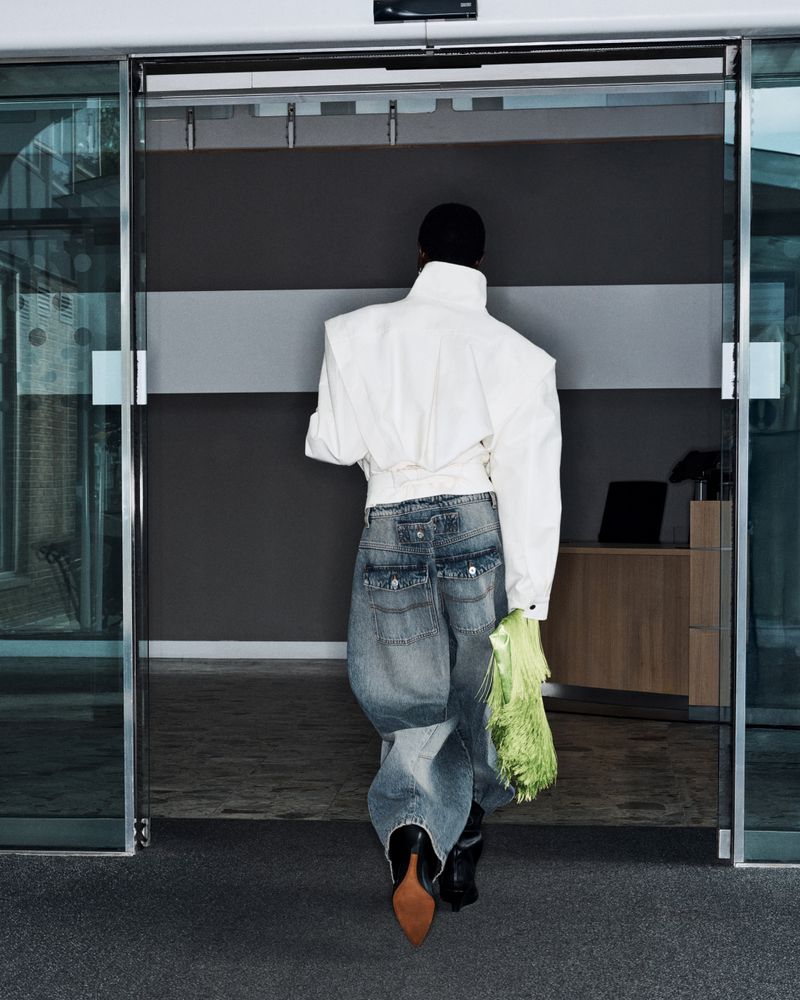Model seen from behind wearing an oversized white structured jacket and baggy denim jeans holding a lime green fringe bag