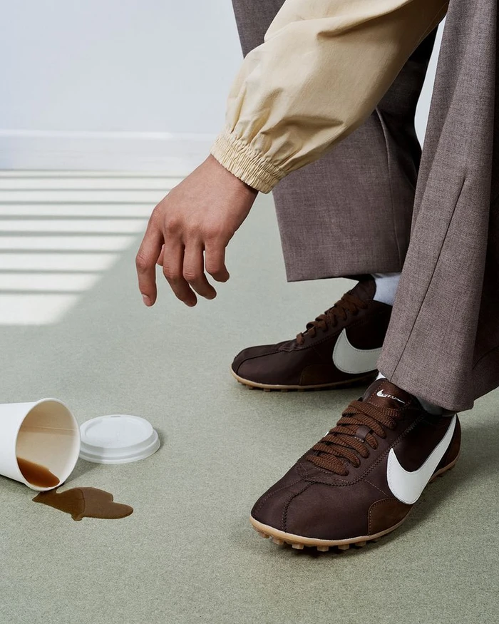 Close-up of a person wearing brown sneakers and trousers standing near a spilled paper coffee cup on a light green floor.