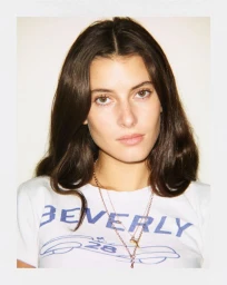 Portrait of a woman with long dark hair wearing a white graphic t-shirt and layered necklaces against a plain white background