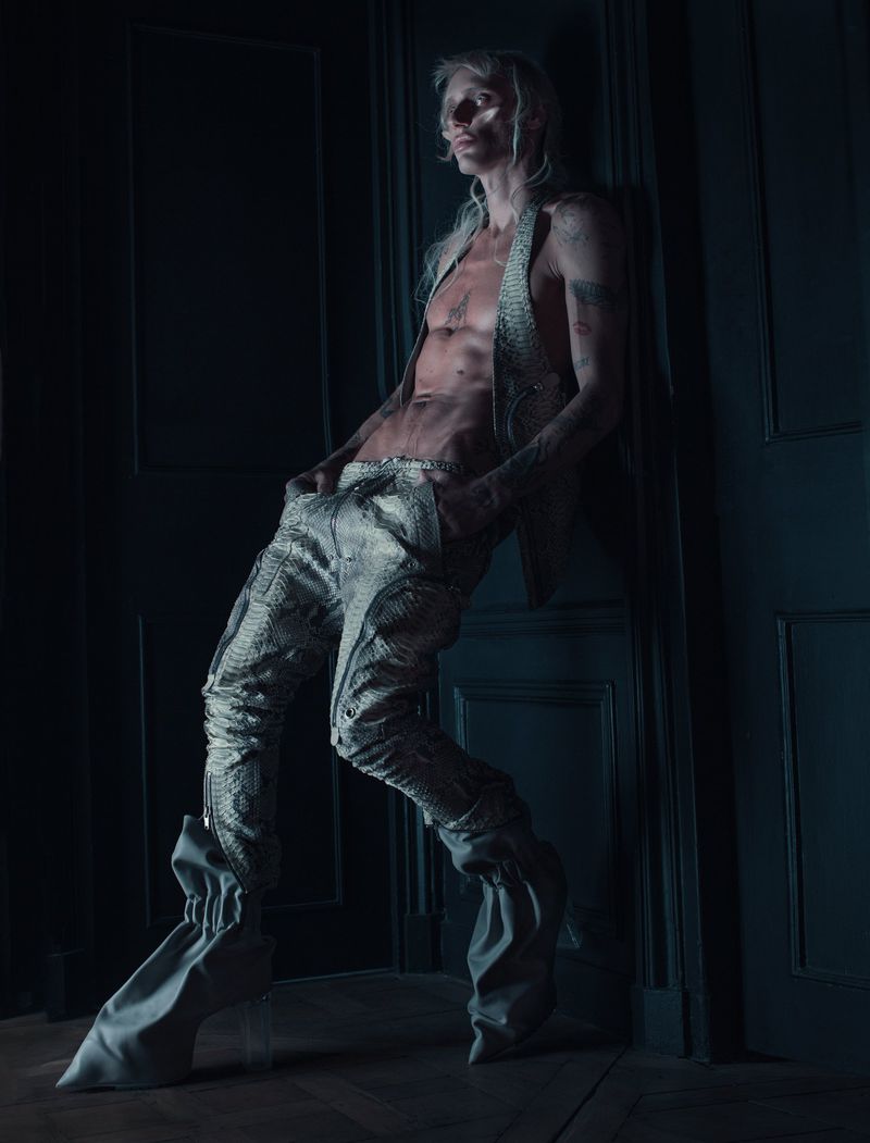 A shirtless male model with blonde hair leaning against a dark panelled wall wearing metallic textured trousers and boots.
