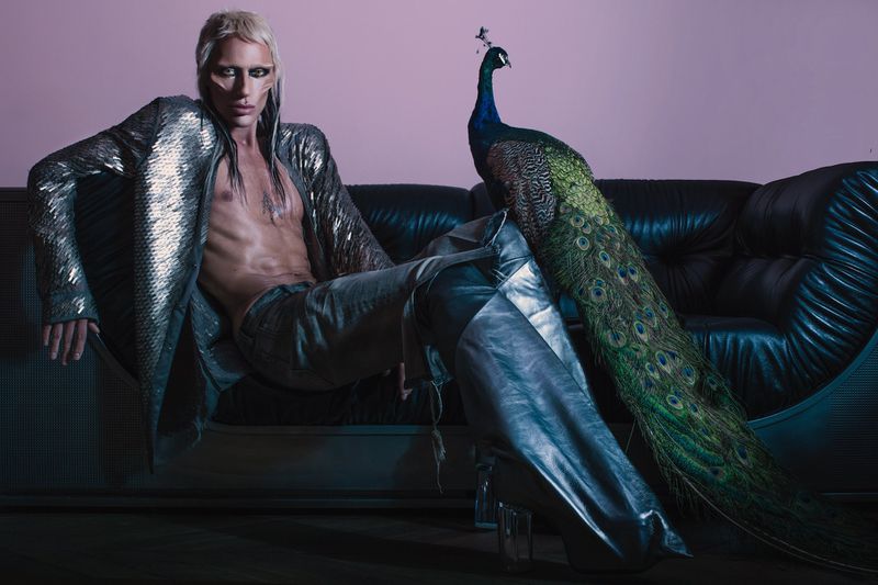 Model with platinum blonde hair and metallic jacket seated on a black leather couch next to a peacock