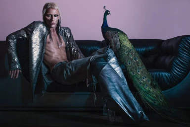 Model with platinum blonde hair and metallic jacket seated on a black leather couch next to a peacock