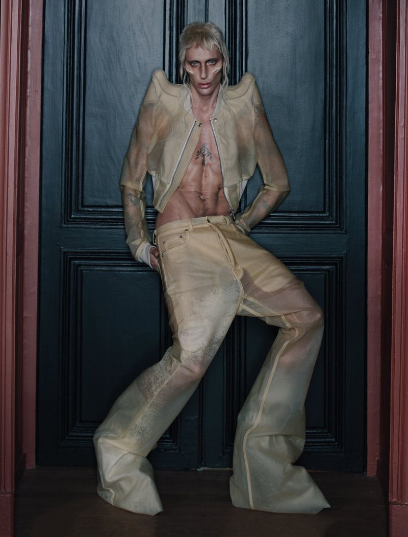 Model with blonde hair posing in a translucent structured jacket and matching trousers against dark double doors