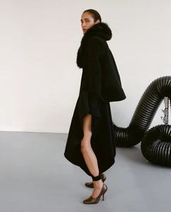 Model Shani Feldman in a black coat with fur collar wearing brown heels against a white studio wall