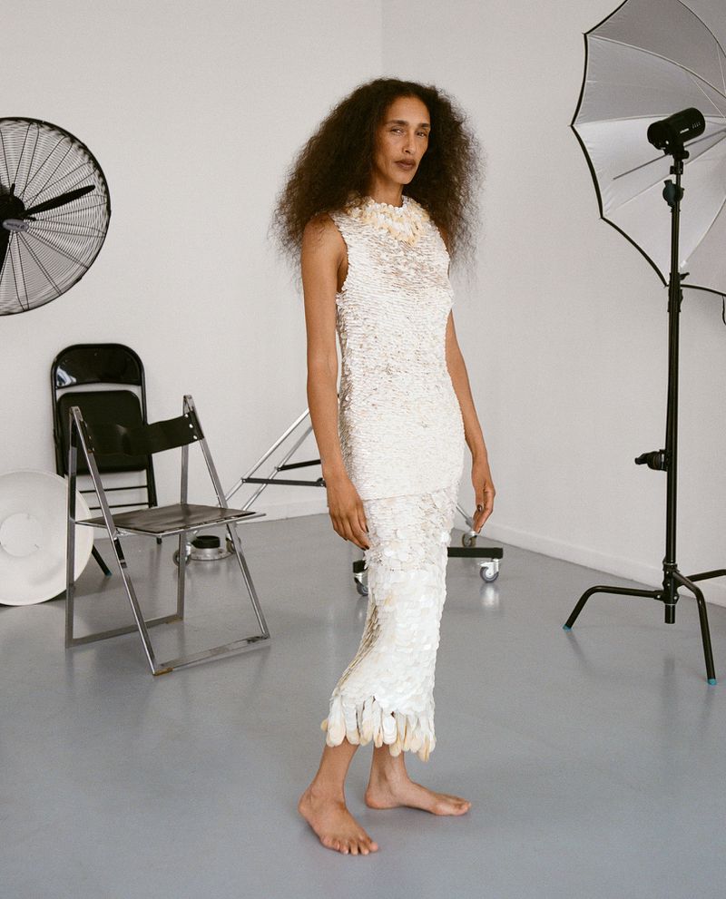 Model with long dark curly hair wearing a white textured dress standing barefoot in a photography studio.