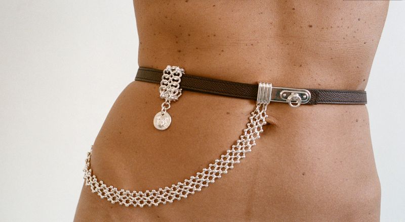 Close-up of a model wearing a brown leather belt with silver chain details against a white background.
