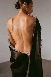 Model Shani Feldman with hair in a bun seen from behind wearing a dark satin coat against a plain neutral studio background