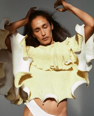 Model Shani Feldman wearing a textured yellow top with white ruffled edges against a grey background.