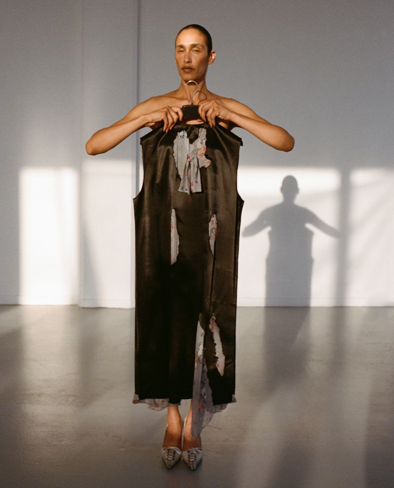Model holding a black dress in front of her body inside a studio setting with a prominent background shadow.