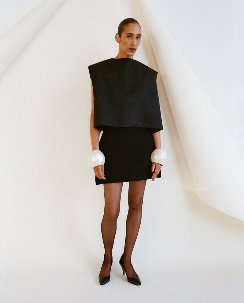 Model wearing a black structured top and skirt with white cuffs against a curved white backdrop