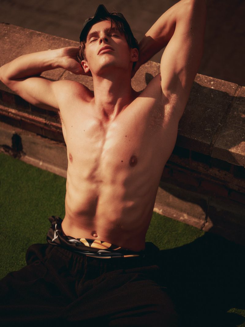 Shirtless male model with hands behind head leaning against a stone ledge under warm natural sunlight.