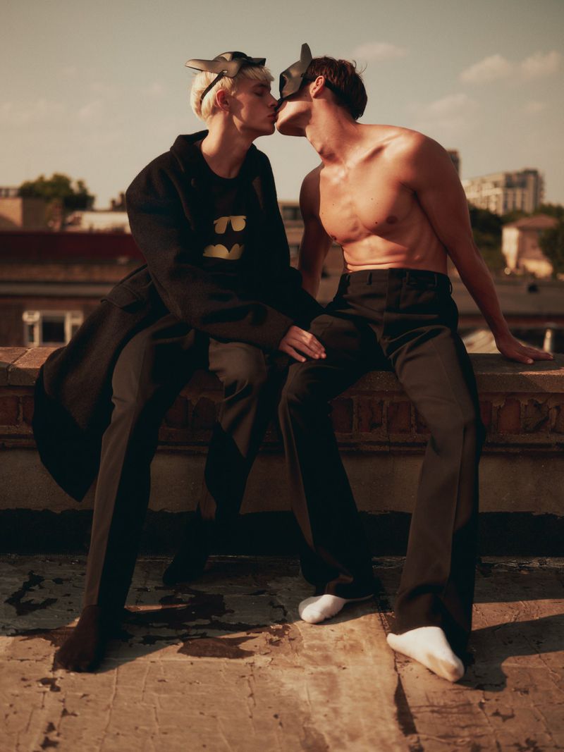 Two men wearing masks kissing while sitting on a rooftop ledge against an urban background