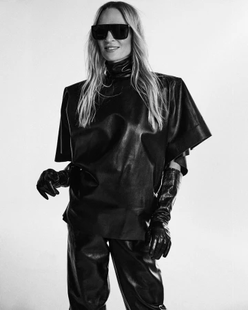 Uma Thurman wearing oversized sunglasses and a black leather top against a plain studio background