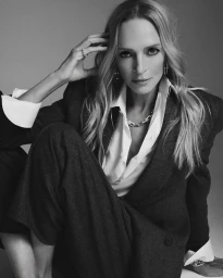 Portrait of Uma Thurman in a dark tailored suit and white shirt, touching her hair against a seamless background.