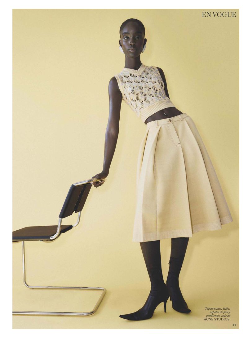 Model leaning against a modern chair in a sleeveless patterned top and pleated skirt against a yellow background